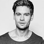 The image features a monochrome portrait of a young man with a relaxed, neutral expression. His hair is styled casually, and he is wearing a simple gray t-shirt. The background is plain, drawing attention to his face and emphasizing his features. In this image, the use of black and white creates a timeless quality, stripping away distractions and encouraging viewers to focus on the subject's expression and the subtle details of his features. This minimalist approach can evoke feelings of introspection or contemplation, inviting the audience to ponder the story or thoughts behind the gaze of the individual. The straightforward style may suggest a modern aesthetic, aligning with themes of authenticity and simplicity in contemporary visual culture.